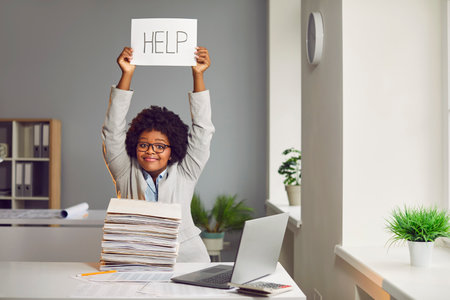 Asking help. Attractive african-american young woman holds piece of paper over her head with inscription help. Dark-skinned girl with pile of folders on her desk. Workplace in office with laptop.の写真素材