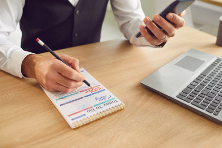 Busy man makes to do list in work planner. Man sitting at office desk checking cellphone, using...
