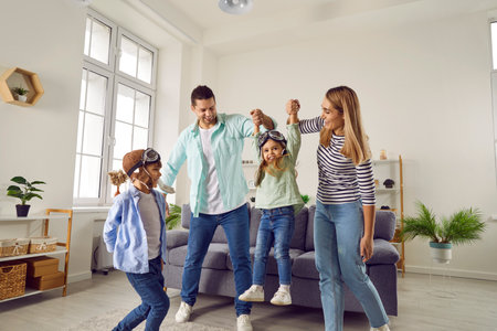 Carefree cheerful parents, son and daughter playing at home. Happy mother and father having fun carrying their daughter wearing goggles by hands like airplane in living roomの写真素材