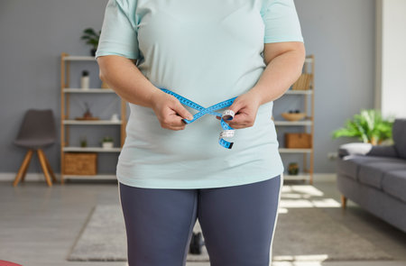 Close up cropped photo of a fat overweight woman wearing sportswear measure the waist with measuring tape standing in the living room at home. Obesity, weight control, diet and slimming concept.の写真素材