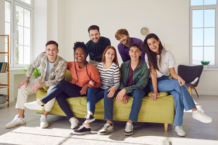 Portrait of a group of happy young diverse friends men and women sitting on sofa in the living room at home looking cheerful at camera enjoying meeting. Friendship, party and home leisure concept.の写真素材