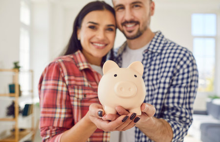 Close up captures a couple hands jointly hold a piggy bank at home. Family smiles radiate joy and togetherness, reflecting shared commitment to saving money for future.の写真素材