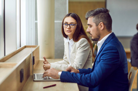 Two people meet and communicate in office. Young woman and man in suits sitting together at table by window, using laptop computer, working on business project and talking about ways to improve itの写真素材