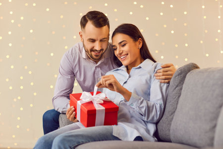Care, congratulations and Valentines day concept. Portrait of a young man congratulating pretty smiling woman on holiday with red present gift box sitting on sofa at home. Happy couple in love.の写真素材