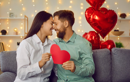 Happy couple holding red paper heart looking at each other. Romantic man and woman celebrating St. Valentine day sitting on background of red heart shaped balloons and glowing lightsの写真素材