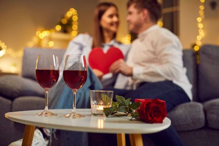 Close up table with two red wine glasses, beautiful rose, candle and gift box. Loving married couple having home date, relaxing on couch, kissing, giving each other Valentine heart cards in backgroundの写真素材