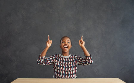 Overjoyed young African American woman sit at desk on black studio background point up at empty...