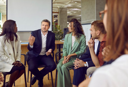 Diverse business team meeting with professional coach. Group of multiethnic people sitting in circle in office interior and listening to mentor sharing his experience, values and leadership mindsetの写真素材