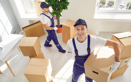 Top view of a young happy smiling employee of moving service in overall standing in the living room of new house holding cardboard box looking cheerful at camera and showing thumb up sign.の写真素材