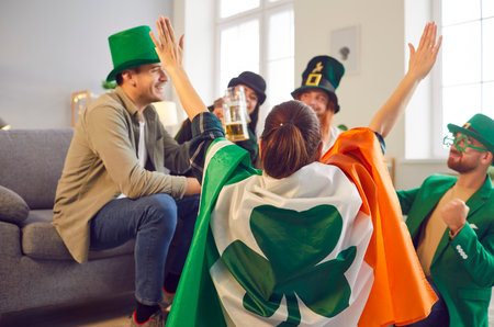 Happy smiling friends celebrating Saint Patricks Day at home sitting in a circle in green clothes and hats. Young men and women talking and having fun on party drinking beer with a flag of Irelandの写真素材