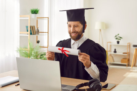 Smiling graduate man having video call showing his diploma. Male student sitting at the desk at home...