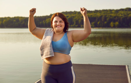Young happy smiling funny fat woman in sportswear do winners gesture clenching fists celebrating success in workout standing outdoors in the park. Healthy lifestyle, sport and fitness concept.の写真素材
