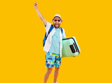 Happy man tourist in summer beach attire, isolated on orange background. With a suitcase and backpack in tow, he exudes the excitement of embarking on a well deserved vacation or holiday.の写真素材
