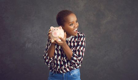 Studio portrait of happy smiling black woman holding pink piggy bank with savings to her ear, listening to sound of coins clinking inside, anticipating spending whole lot of money on fulfilling dreamsの写真素材