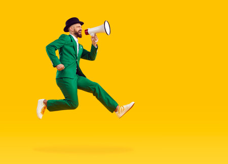 Side profile view full body length man in green suit and hat jumping up, flying on yellow copy space background, screaming through megaphone, making announcement, advertising Saint Patricks Day partyの写真素材