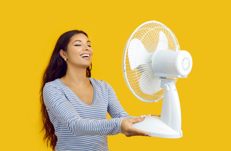 Summer heat. Joyful woman enjoys flow of air from an electric fan isolated on yellow background....