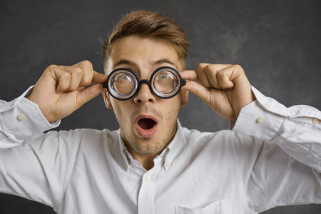 Close up portrait of a crazy surprised young man in glasses with magnifying lenses. Caucasian man in a white shirt with a shocked expression on a background of gray concrete.の写真素材