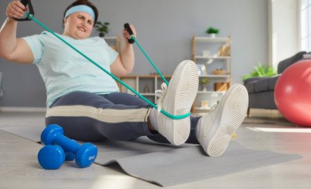 Fat obese woman doing stretching exercises with rubber band sitting on the floor at home. Plus size young girl doing domestic workout. Sport, gym fitness and body positive concept.の写真素材