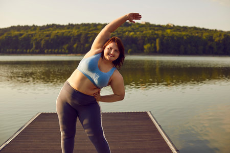Happy fat young woman in sportswear doing stretching sport exercises in the summer city park on...