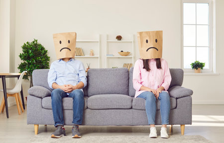 Faceless man and woman sitting on opposite sides of sofa with paper bags on head with upset faces ignoring each other. Stressed married couple sitting separately at home. Quarrel and divorce concept.の写真素材