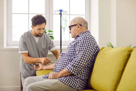 Male caregiver preparing elderly patient for IV therapy at home. Senior man sitting on sofa receiving intravenous treatment or vitamin therapy at home. Elderly people health care and medical supportの写真素材