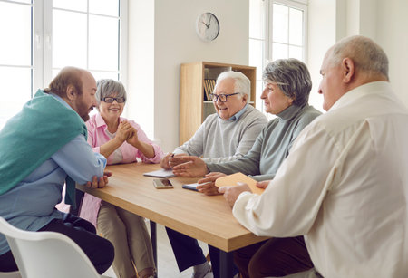 Group of elderly people are talking and having good time together sitting at table at home. Cheerful elderly people talking while sitting at table in modern retirement home. Senior social life conceptの写真素材