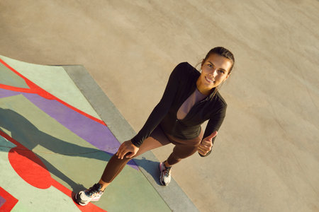 Outdoor portrait of sporty woman giving thumbs up. High angle view happy beautiful young girl in sports clothes standing on urban street or skatepark concrete looking up doing like gesture and smilingの写真素材