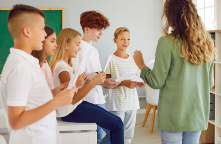 Group of happy smiling junior school children boys and girls standing in the classroom holding white sheets in hands with their young female teacher and singing in choir during music lesson.の写真素材