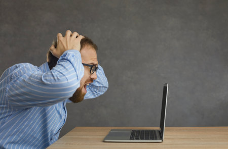 Side profile view man sitting at desk, holding head in hands, looking at laptop screen in despair....
