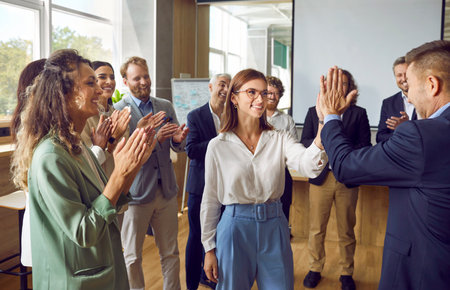 Team of happy business people celebrating success together. Group of men and women having fun at work in office. Smiling female worker high fives colleague while others clap hands. Teamwork conceptの写真素材