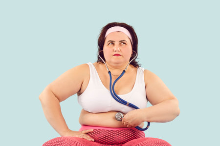 Obese large chubby overweight woman in sports clothes holds stethoscope on skin of big fat belly,...