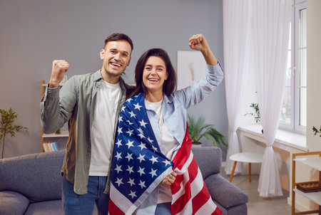 Young happy cheerful couple patriots man and woman with American flag standing in living room at...