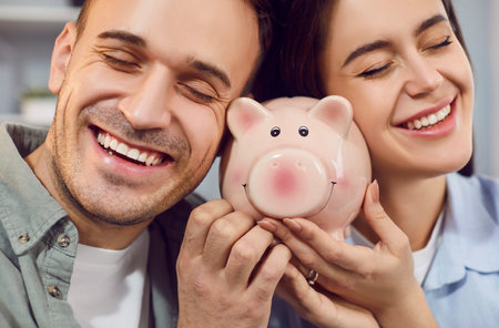Close up of happy young family holding piggy bank indoors to save money for future. Married couple planning saving their budget together. Investments and financial freedom concept.の写真素材