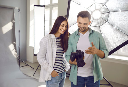 Portrait of a young smiling professional photographer showing photos to a girl model on digital camera standing in production studio with light equipment and discussing photo session.の写真素材