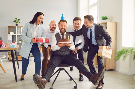 Cheerful happy man celebrating his birthday in office with his colleagues. Man in party hat is sitting in office chair holding birthday cake while his satisfied colleagues surround him with gifts.の写真素材