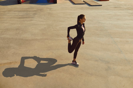 Young woman in sports clothes having outdoor fitness workout on city street, standing on grey concrete sportsground surface, getting ready to start jogging and doing warming up leg stretching exerciseの写真素材