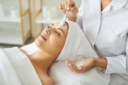 Cosmetologist applying moisturizing mask with brush to a young relaxed smiling young woman in spa salon. Close up portrait of a girl client receiving beauty procedure from beautician in beauty clinicの写真素材