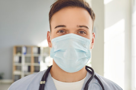 Close-up portrait of young man doctor wearing medical mask with stethoscope looking at camera ready...