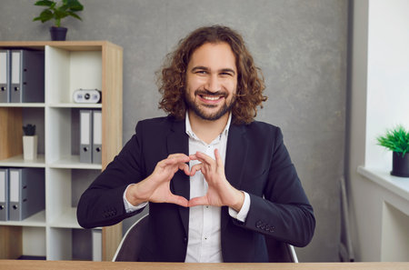 Portrait of a smiling happy man with beard and long hair wearing business clothes standing in office doing heart symbol shape with hands as a sign of love looking cheerful at the camera.の写真素材