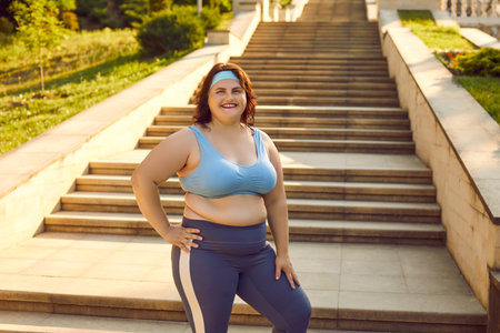 Overweight young fat woman wearing sportswear standing in summer park drinking a water after doing fit exercises outdoors. Workout sport, fitness in nature and body positive concept.の写真素材