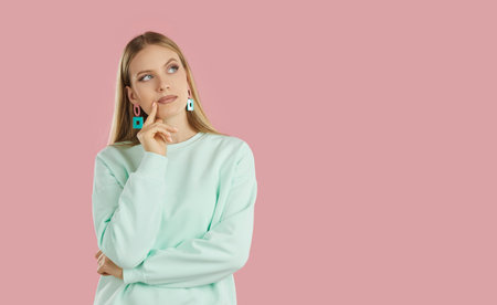 Thoughtful bewildered serious blondy girl in mint sweatshirt with geometric earrings looking up on...