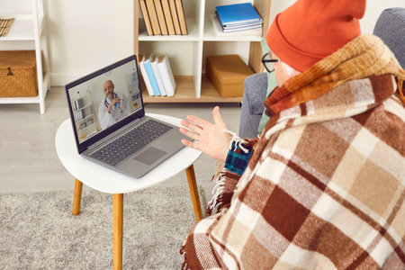 Sick senior man talking about the symptoms of his illness having online video call with a male confident doctor using laptop at home. Telemedicine and patient consultation online concept.の写真素材