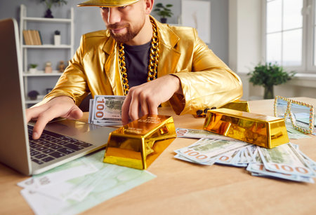Funny happy Caucasian man in golden suit sitting at desk with money bills at home with laptop working or doing online shopping. Male person with cash and gold on table. Investment and success conceptの写真素材