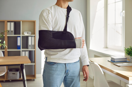 Close-up cropped photo of a mans broken arm in a plaster cast standing at home or at office on rehabilitation after injury. Patient wearing sling or bandage. Recovery and rehab conceptの写真素材