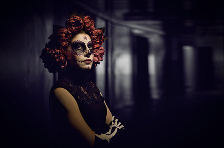 Portrait of woman dressed for Halloween or Day of the Dead. Lady with rose wreath and Catrina skull makeup standing on dark background inside dark building looking at camera with unfriendly expressionの写真素材