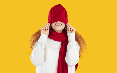 Funny teenage girl in scarf behaves cheerfully and playfully, pulling red warm hat on her eyes. Cheerful curly red-haired girl hides her eyes behind hat isolated on yellow background. Web banner.の写真素材