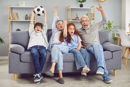 Excited happy family of grandfather, grandmother and children football fans watching sport match on tv supporting their favorite team together sitting on sofa at home with hands up and ball.の写真素材