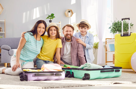 Portrait of happy family of four packing bag for vacation trip. Father, mother with children sitting on the floor at home looking cheerful at camera. Family summer holiday preparations.の写真素材