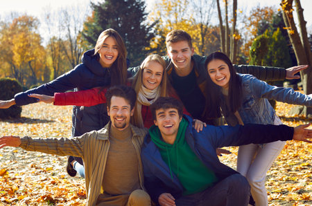 Smiling friends in jackets and scarves pose for photo, hugging and spreading their arms out to the sides. Group portrait of happy young friends strolling through the park on clear sunny autumn day.の写真素材