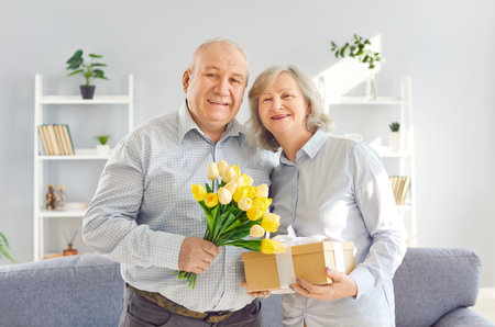 Happy smiling senior couple standing in living room at home with tulips flowers bouquet and present gift box celebrating marriage anniversary holiday and looking cheerful at camera.の写真素材